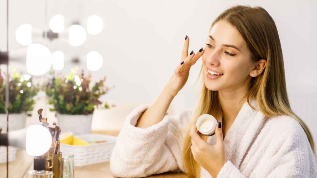 Young woman applying face cream skincare moisturizer at vanity mirror.