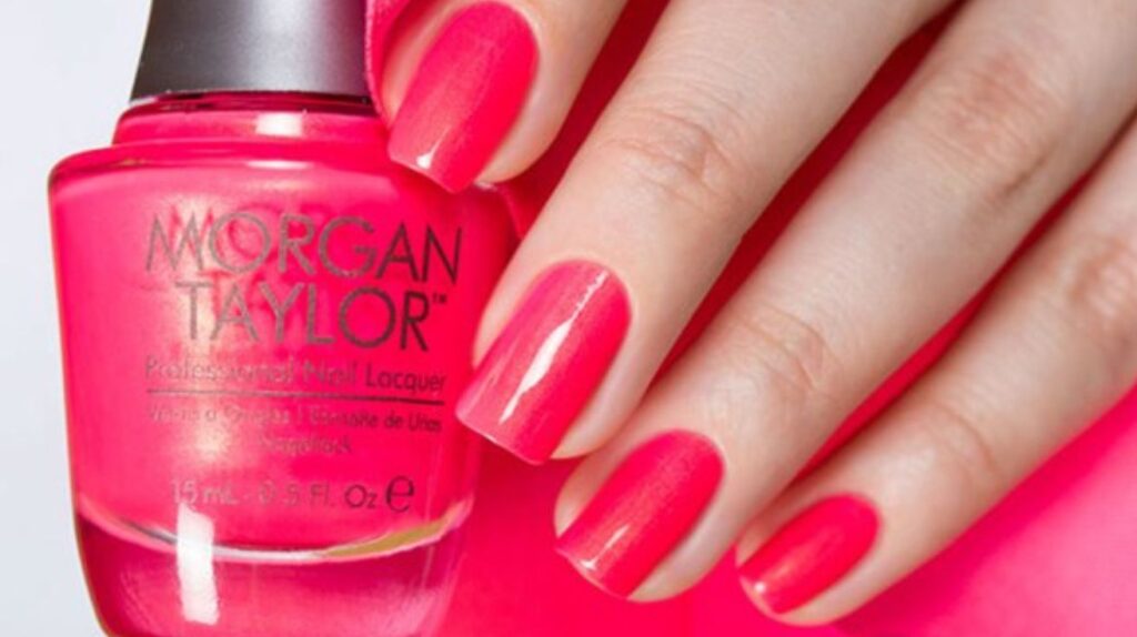 Glossy coral nail polish on manicured nails with soft pastel pink floral background