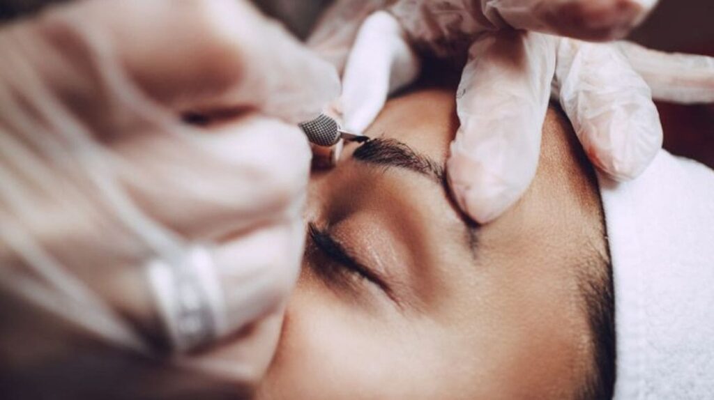 A technician in white gloves performing a microblading procedure on a client's eyebrows, with the client lying back with their eyes closed.