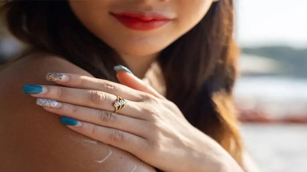 a woman applying sunscreen on her shoulder, wearing a gold ring and colorful nail polish, on a beach.